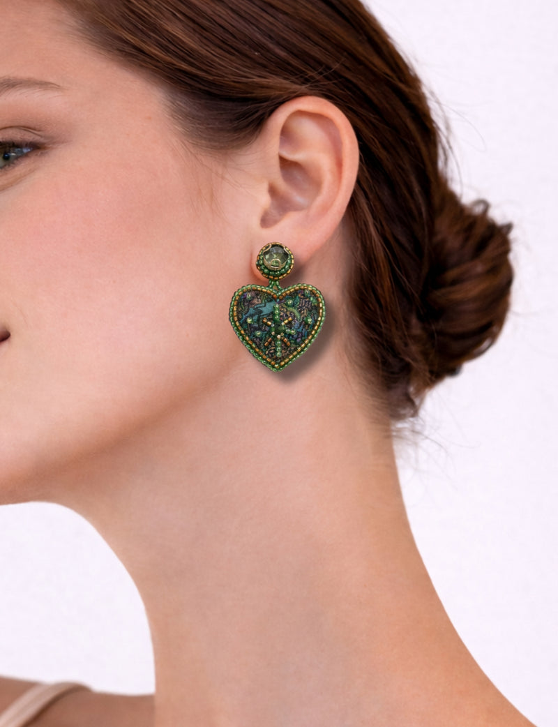 Green heart-shaped drop earring with bead and sequin embellishments worn by a woman with brown hair in a bun, side view