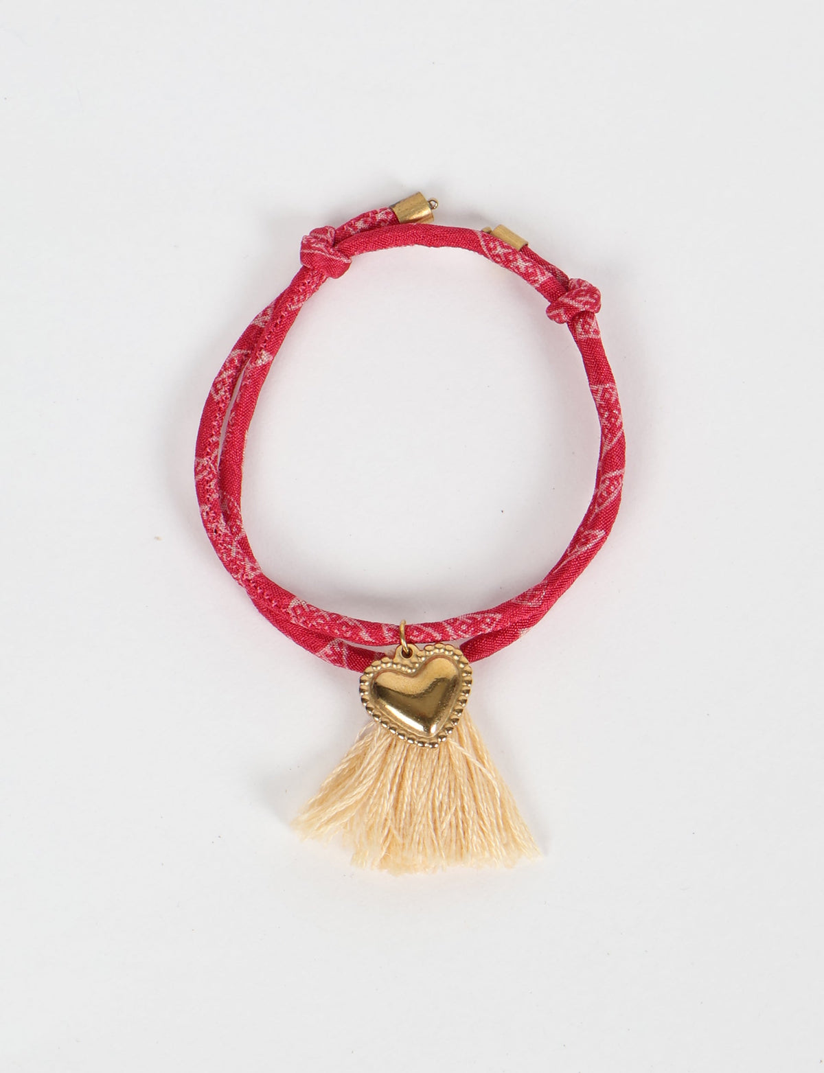 Red patterned rope bracelet with gold heart charm and cream tassels, shown flat on white background