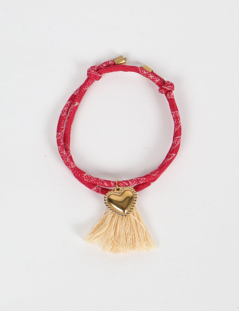 Red patterned rope bracelet with gold heart charm and cream tassels, shown flat on white background