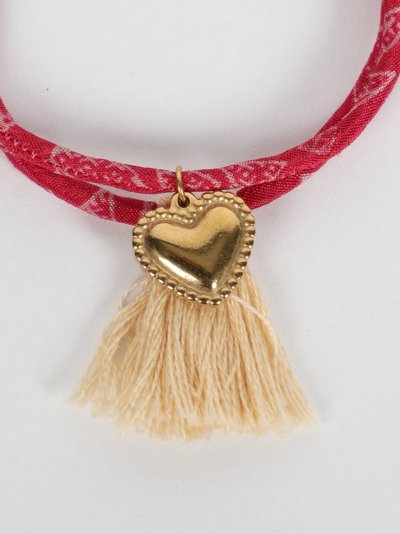 Red patterned rope bracelet with gold heart charm and beige tassels, close-up front view on white background