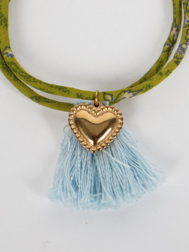Green abstract patterned rope bracelet with gold heart charm and light blue tassels, flat lay on white background