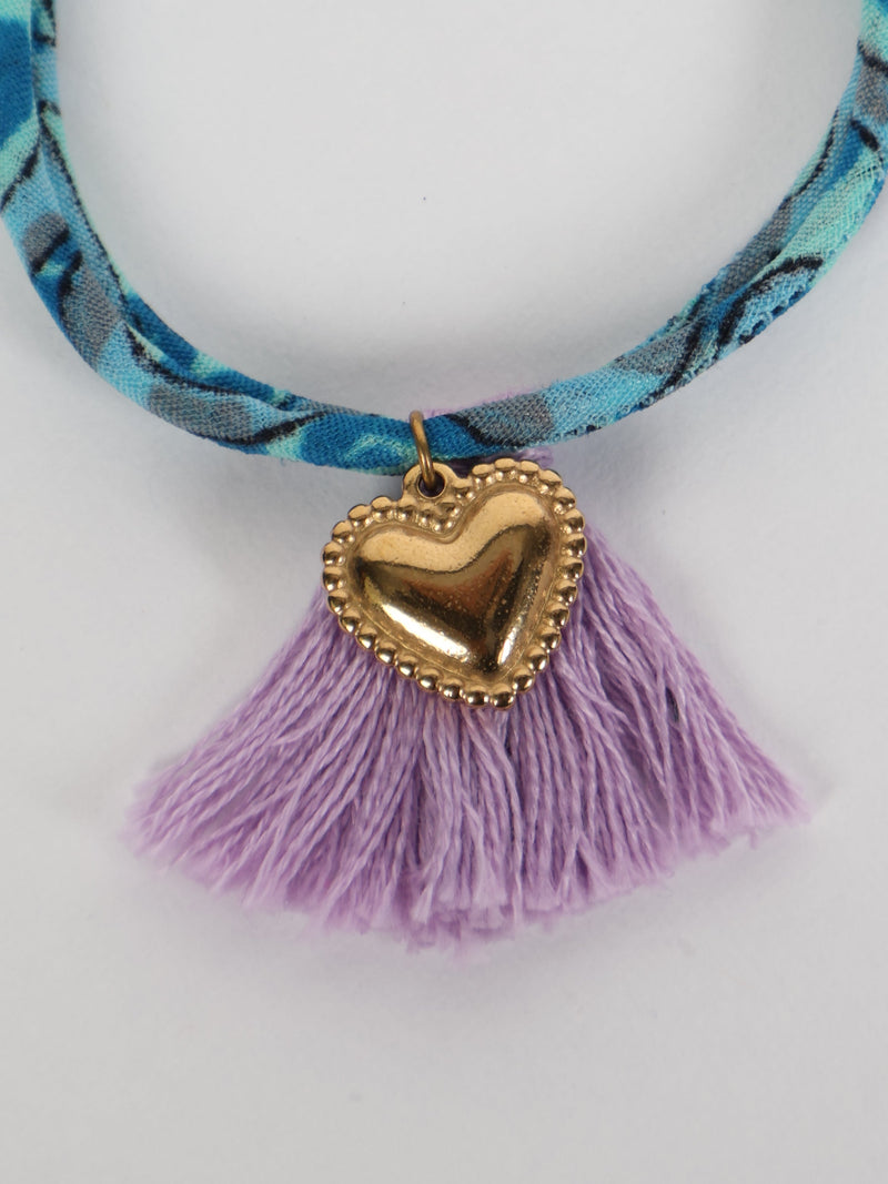 Blue patterned rope bracelet with gold heart charm and light purple tassels, shown in close-up front view