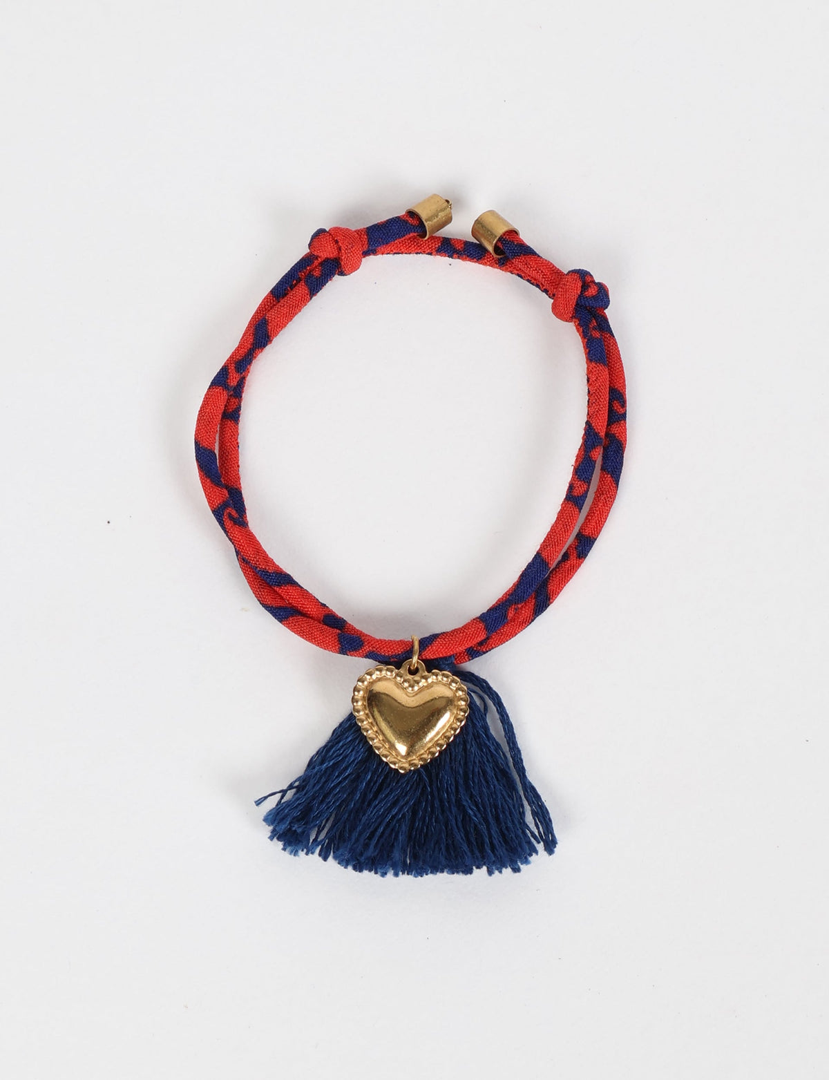 Red and navy abstract patterned rope bracelet with gold heart charm and navy blue tassels, flat lay on white background