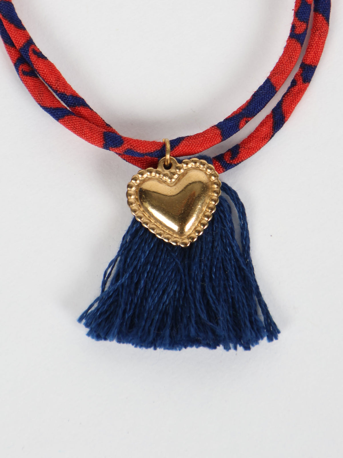 Red and blue patterned rope bracelet with gold heart charm and dark blue tassels, close-up front view