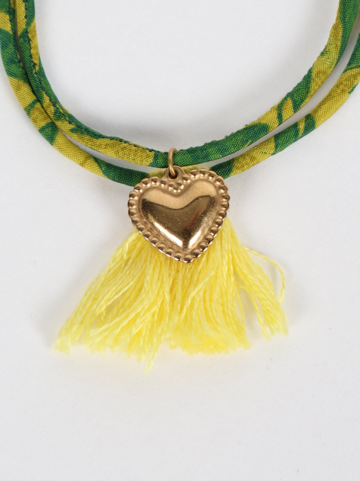 Green and yellow abstract patterned rope bracelet with gold heart charm and yellow tassels, close-up front view
