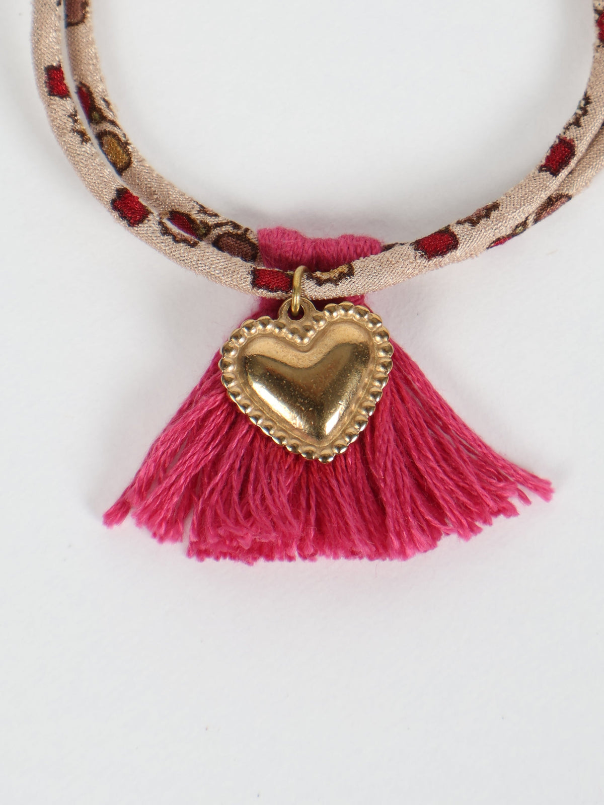 Beige rope bracelet with red and brown abstract pattern, pink tassels, and gold heart charm, flat lay on white background