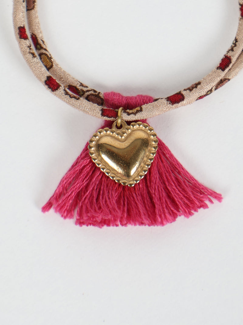 Beige rope bracelet with red and brown abstract pattern, pink tassels, and gold heart charm, flat lay on white background