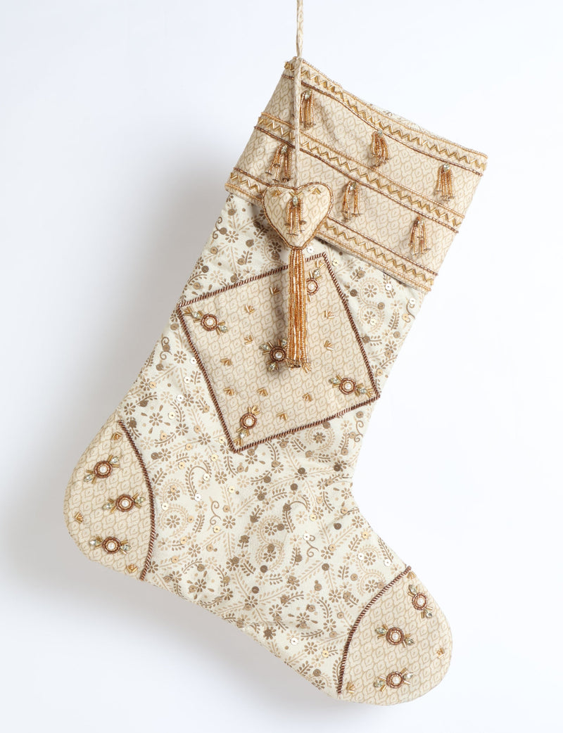 Beige patchwork Christmas stocking with leaf patterns and gold bead tassel embellishments hanging against white background