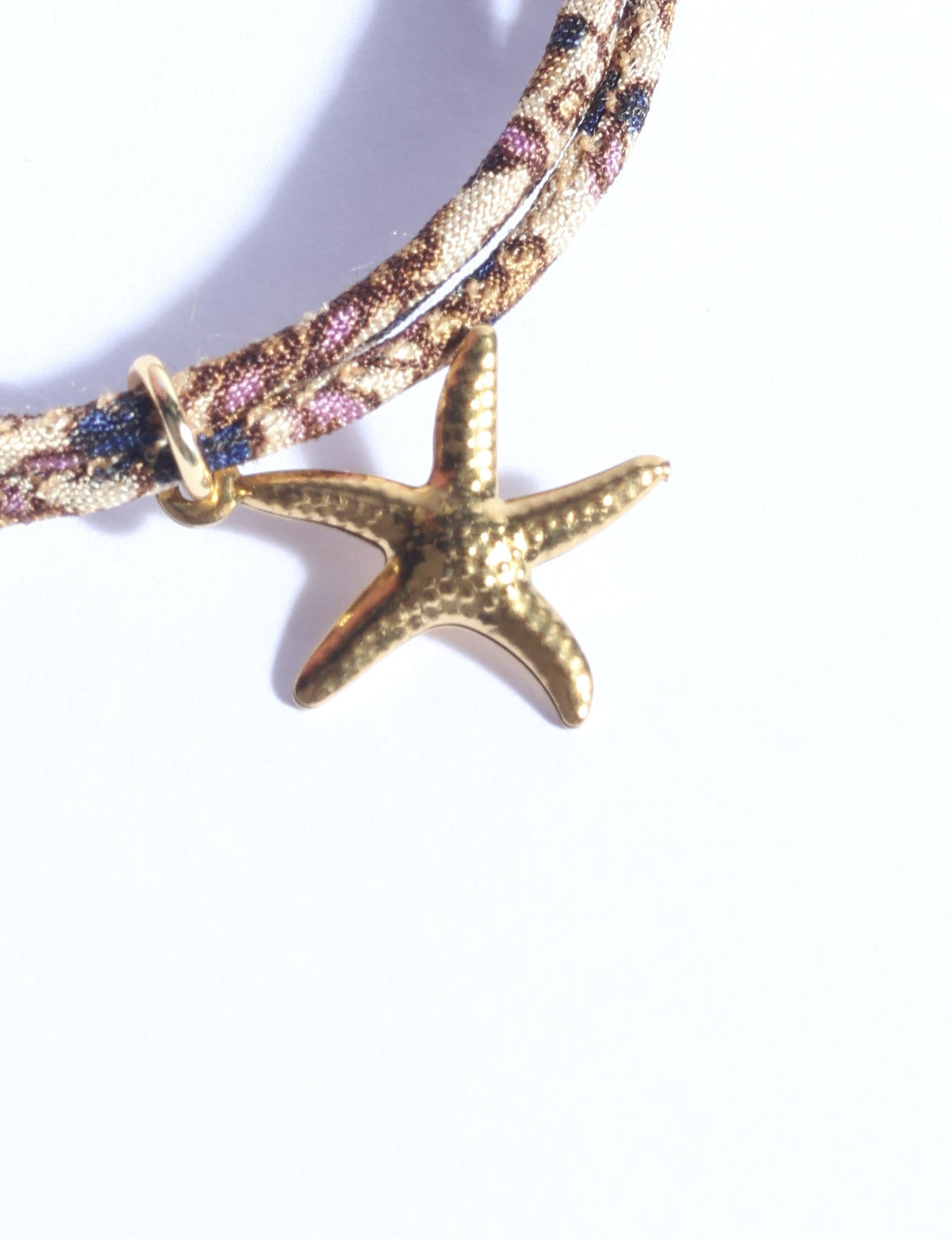 Close-up of a cord bracelet with a gold starfish charm and a patterned fabric cord in beige, brown, blue, and purple tones