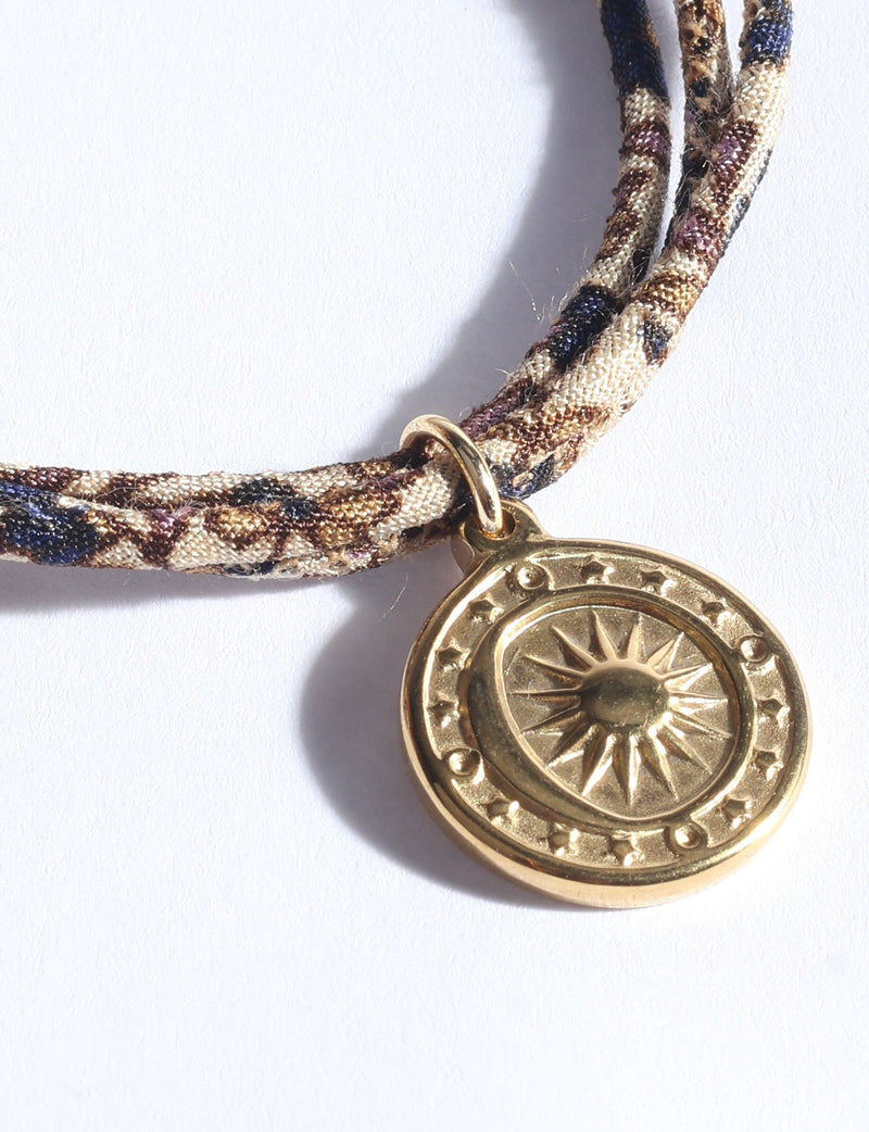 Beige abstract patterned cord bracelet with a round gold-tone sun medallion charm, shown in close-up flat lay