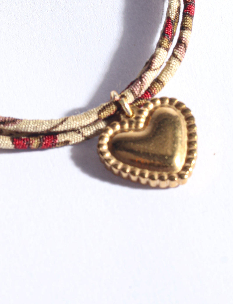 Beige cord bracelet with red and brown abstract pattern and gold-tone heart-shaped charm, close-up view on white background