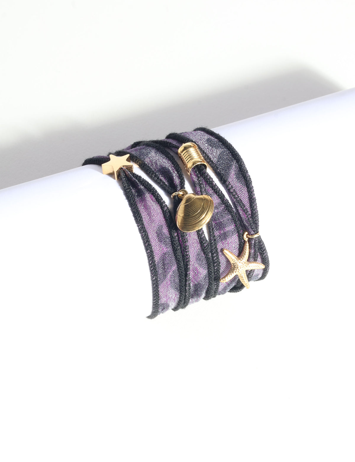 Purple and black wrap-around fabric bracelet with gold star, shell, and starfish charms, shown flat on white surface