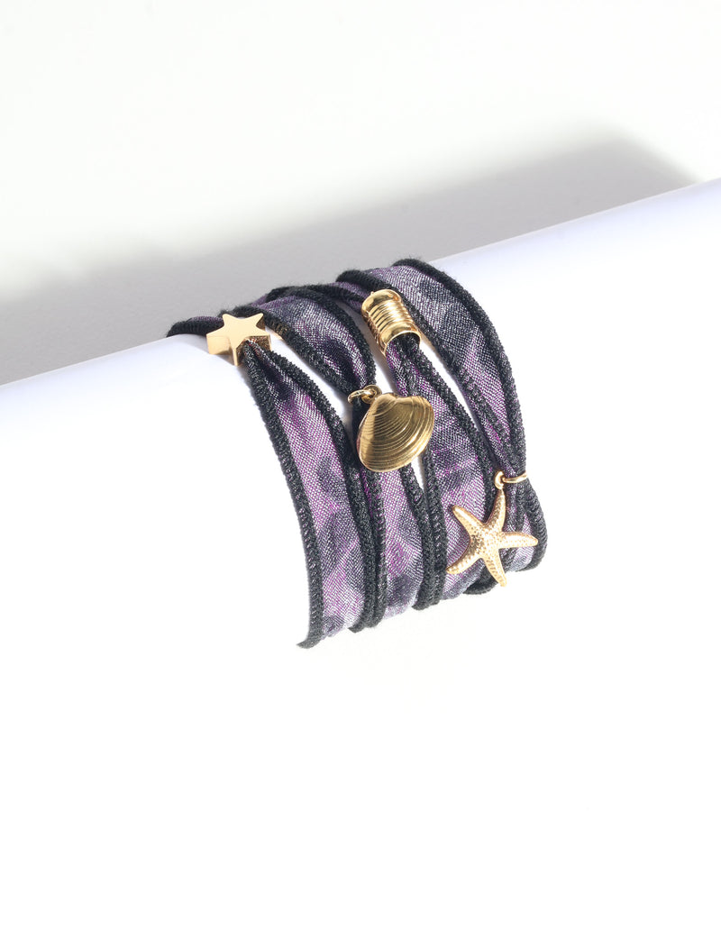 Purple and black wrap-around fabric bracelet with gold star, shell, and starfish charms, shown flat on white surface