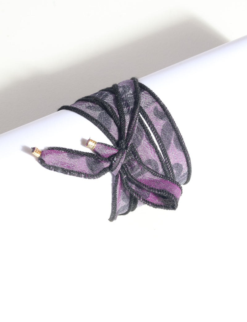 Purple and black wrap-around fabric bracelet with gold-tone charm ends, shown tied on a white cylindrical display