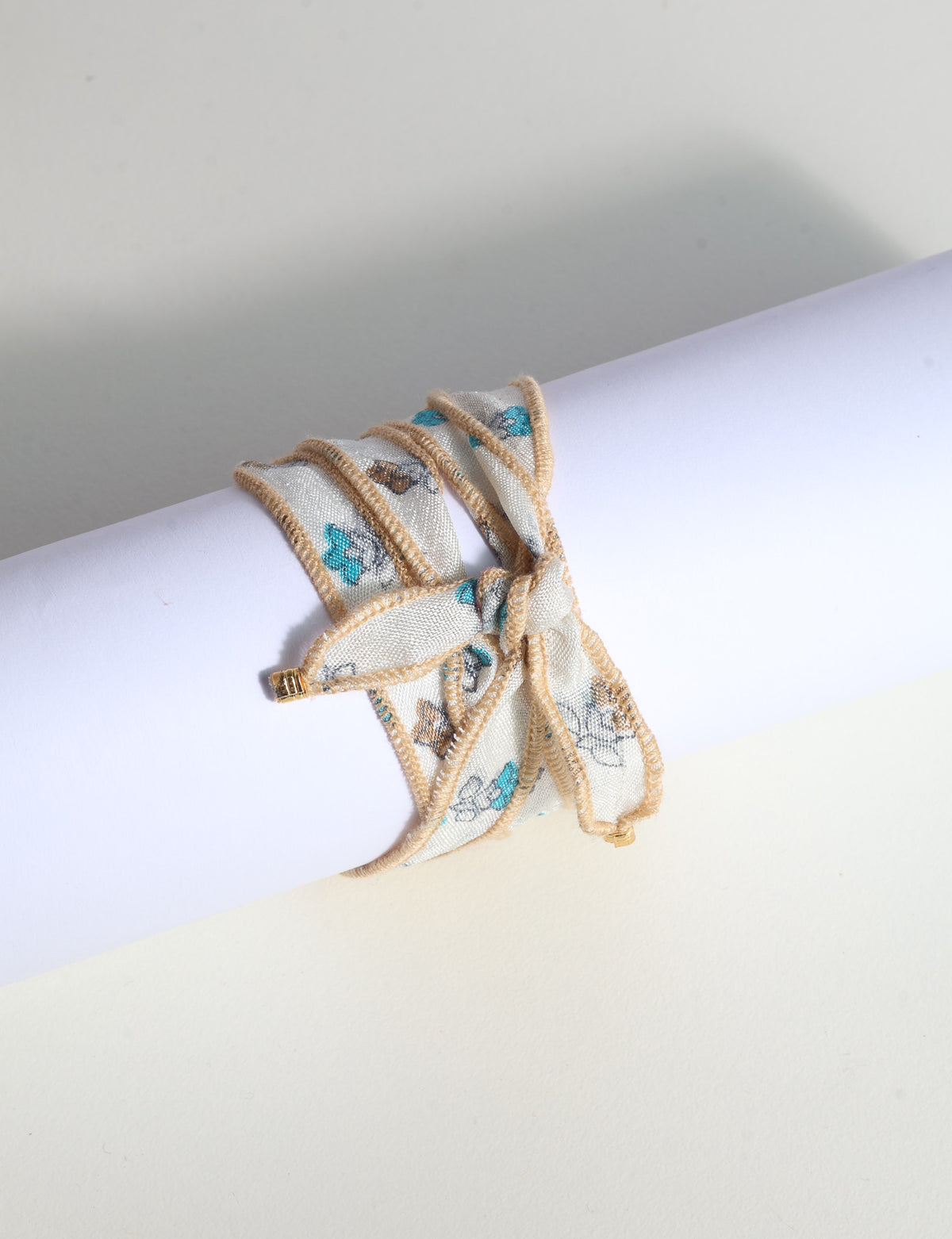 Wrap-around fabric bracelet with beige edges and blue and brown floral print, tied in a bow on white background