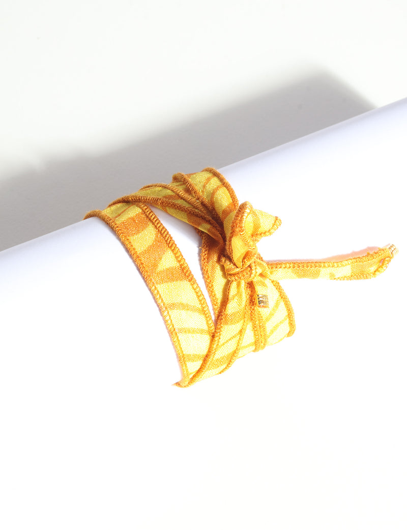 Orange abstract patterned fabric wrap-around bracelet tied in a bow with visible gold charm, shown on white background