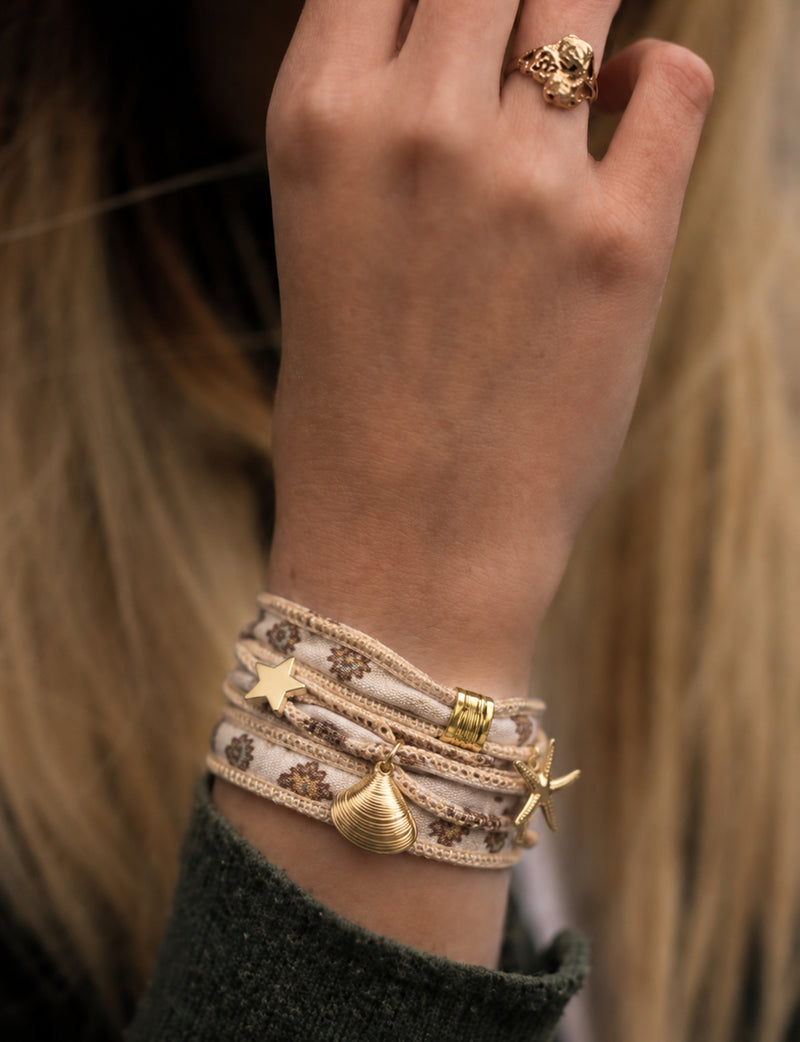 Beige wrap-around fabric bracelet with brown floral print and gold-tone star, shell, and starfish charms worn on wrist