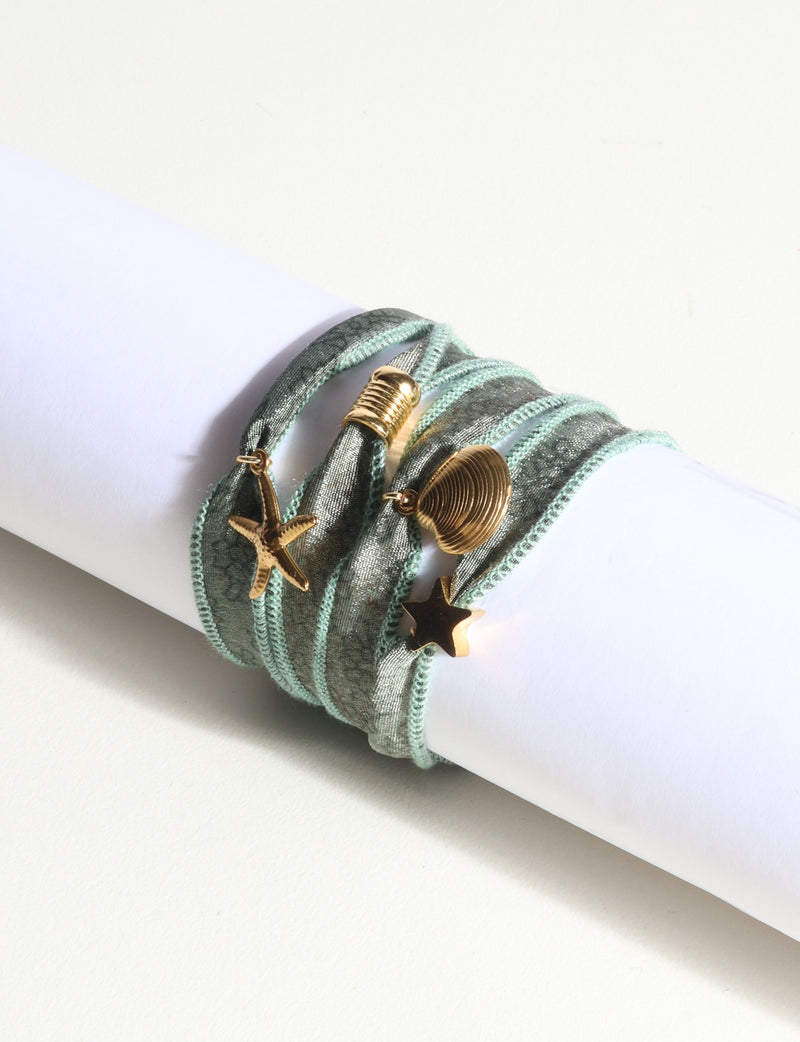Green wrap-around fabric bracelet with gold starfish, shell, and star charms, shown on a white cylindrical display