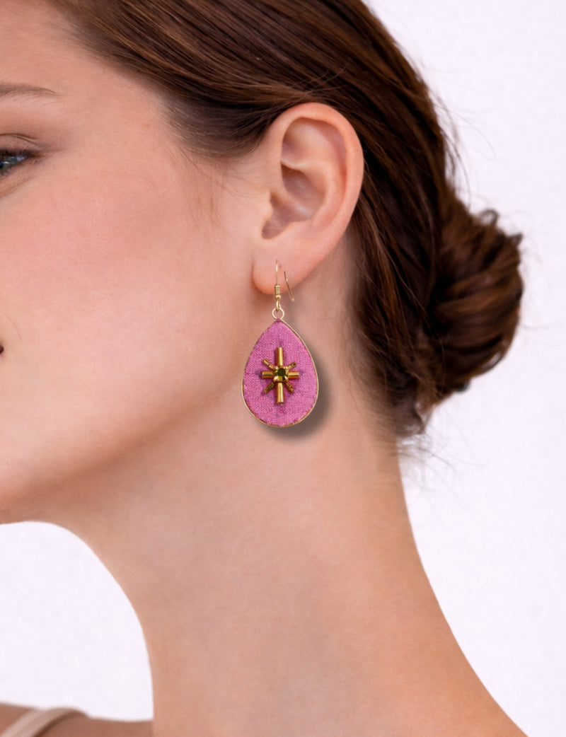 Pink teardrop-shaped earrings with gold-tone cross embellishment worn on a woman's ear, side view close-up