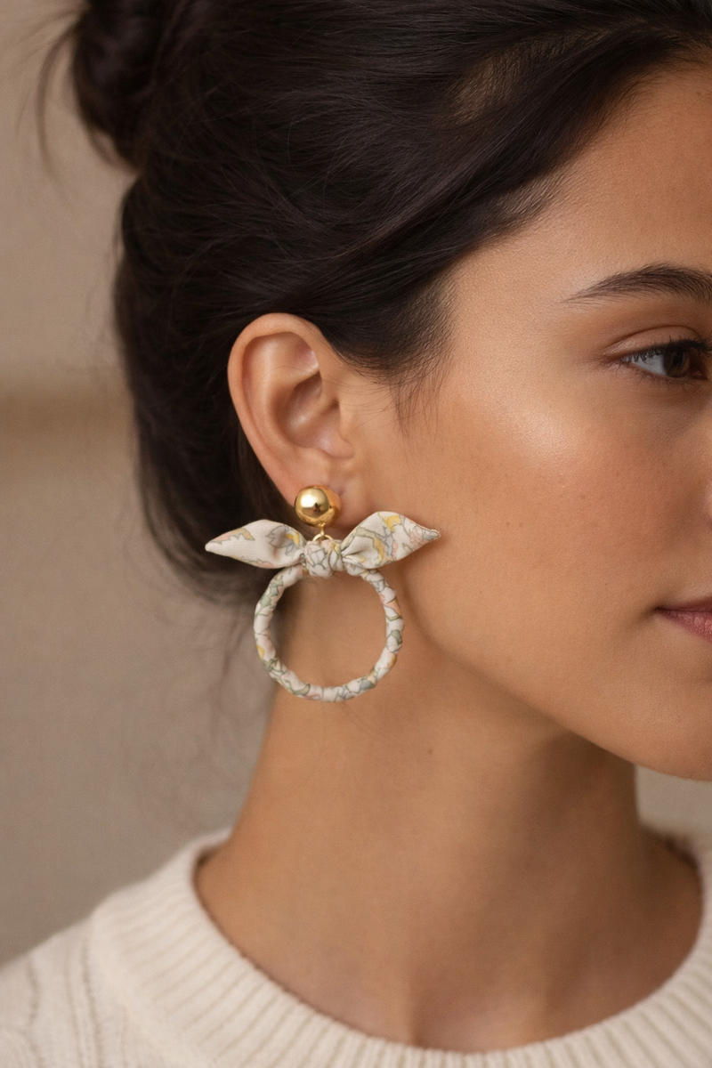 Beige tropical fabric bow earrings with gold ball studs worn by a woman with dark hair in a cream knit sweater