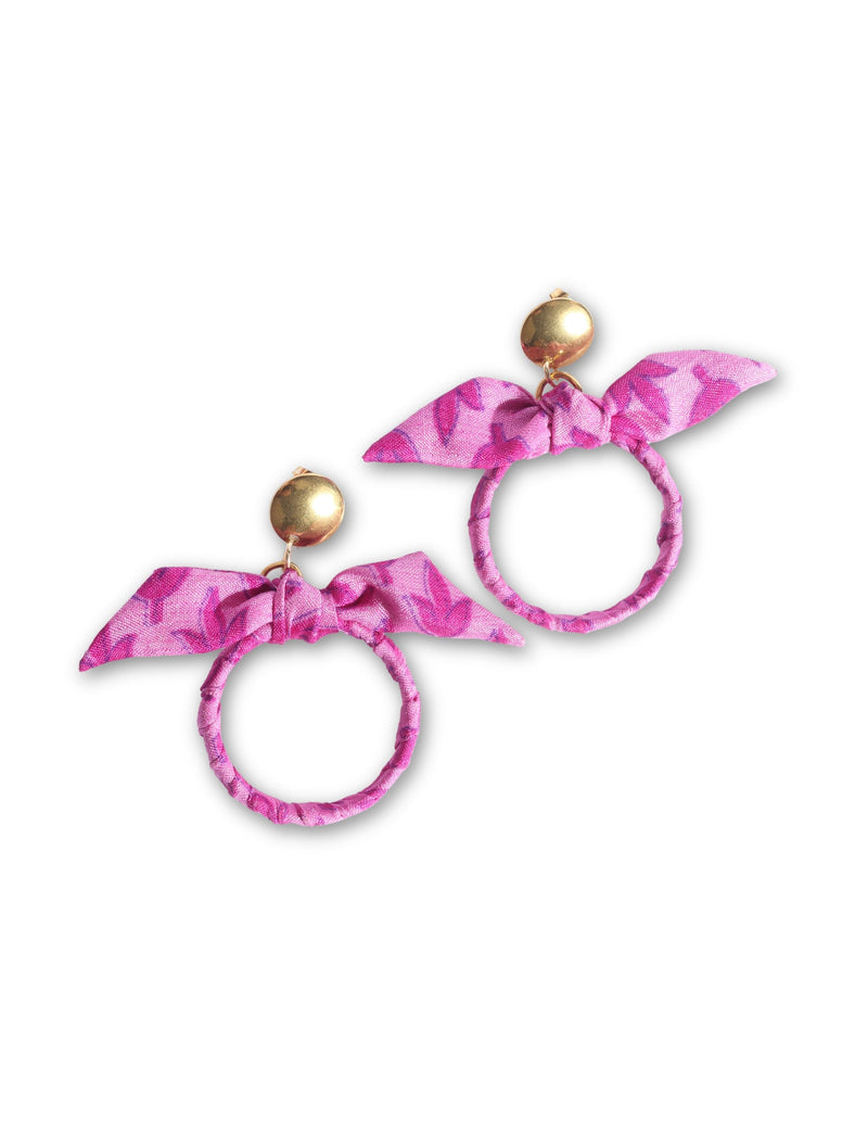 Pink fabric bow earrings with leaf pattern, gold round stud tops, and fabric-wrapped circular hoops, flat lay on white background