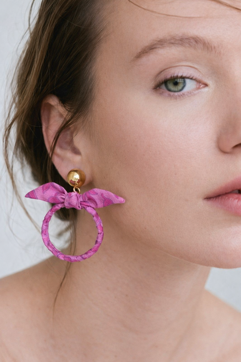 Close-up of a model wearing pink fabric bow earrings with a gold round stud and leaf pattern on the fabric