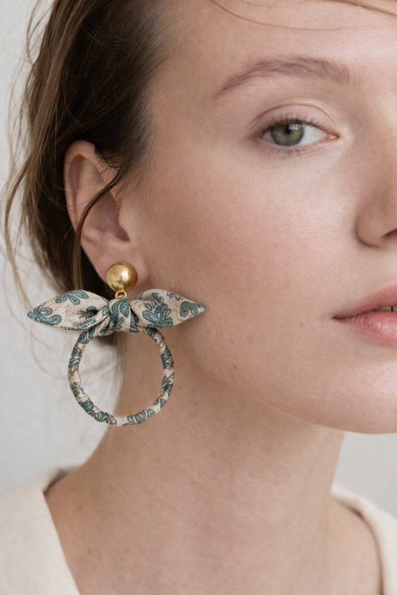 Beige tropical fabric bow earrings with green leaf pattern and gold round stud, worn by model in close-up side view