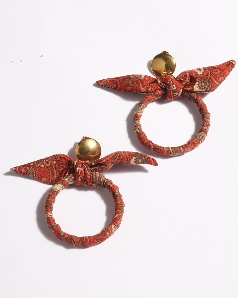 Pair of orange tropical bow earrings with gold round studs and fabric-wrapped circular hoops, flat lay on white background