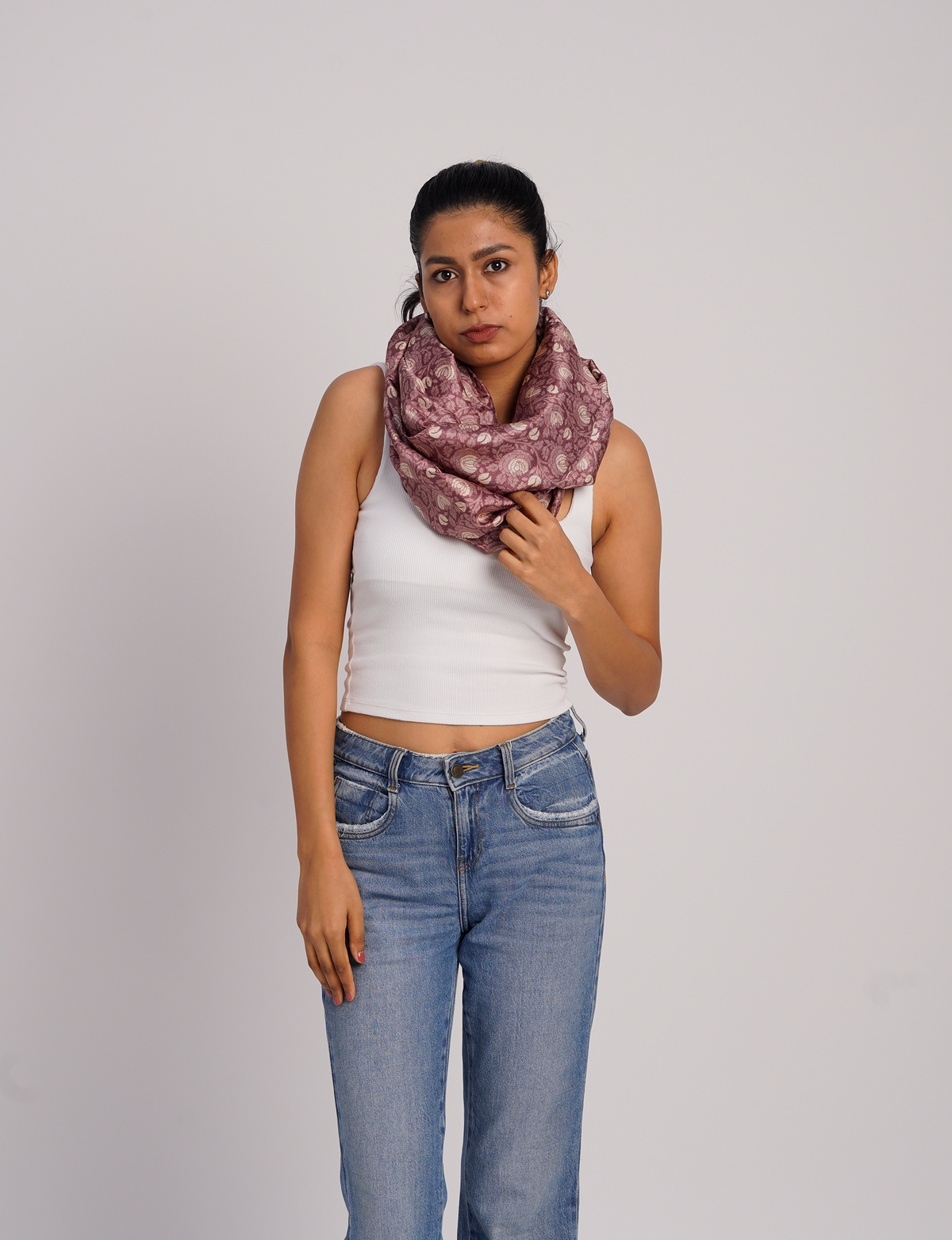 Woman wearing a pink abstract patterned loop scarf, white sleeveless top, and blue jeans, front view on plain background