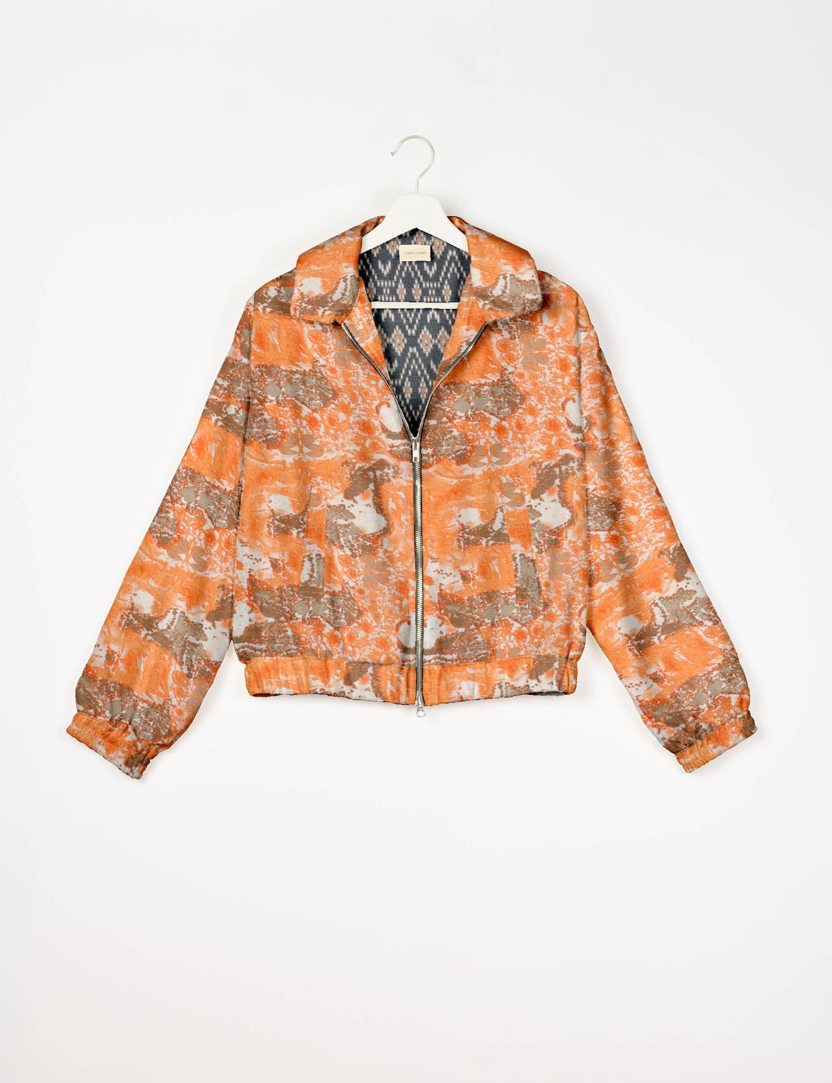 Orange abstract patterned bomber jacket with front zipper and elastic cuffs, shown hanging on a white hanger