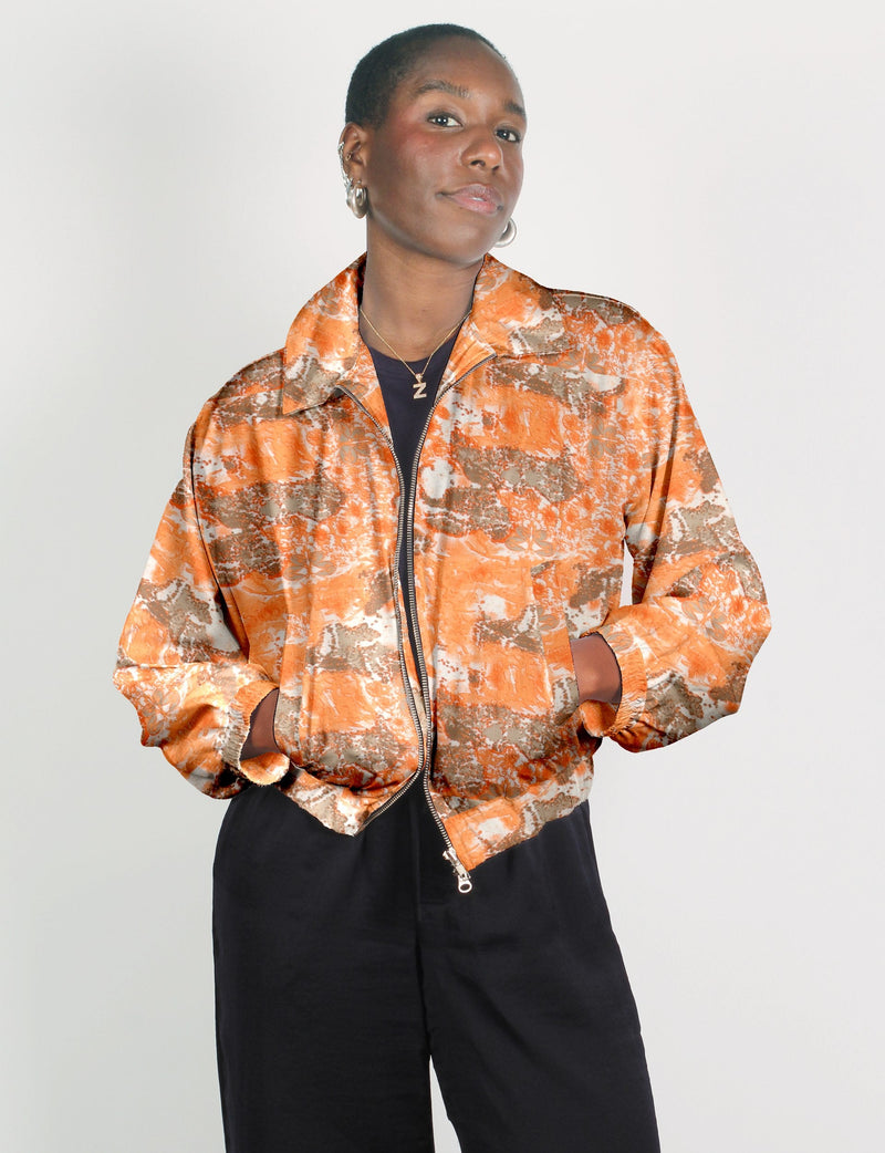 Model wearing an orange and beige abstract patterned bomber jacket with front zipper and elastic cuffs, paired with black pants