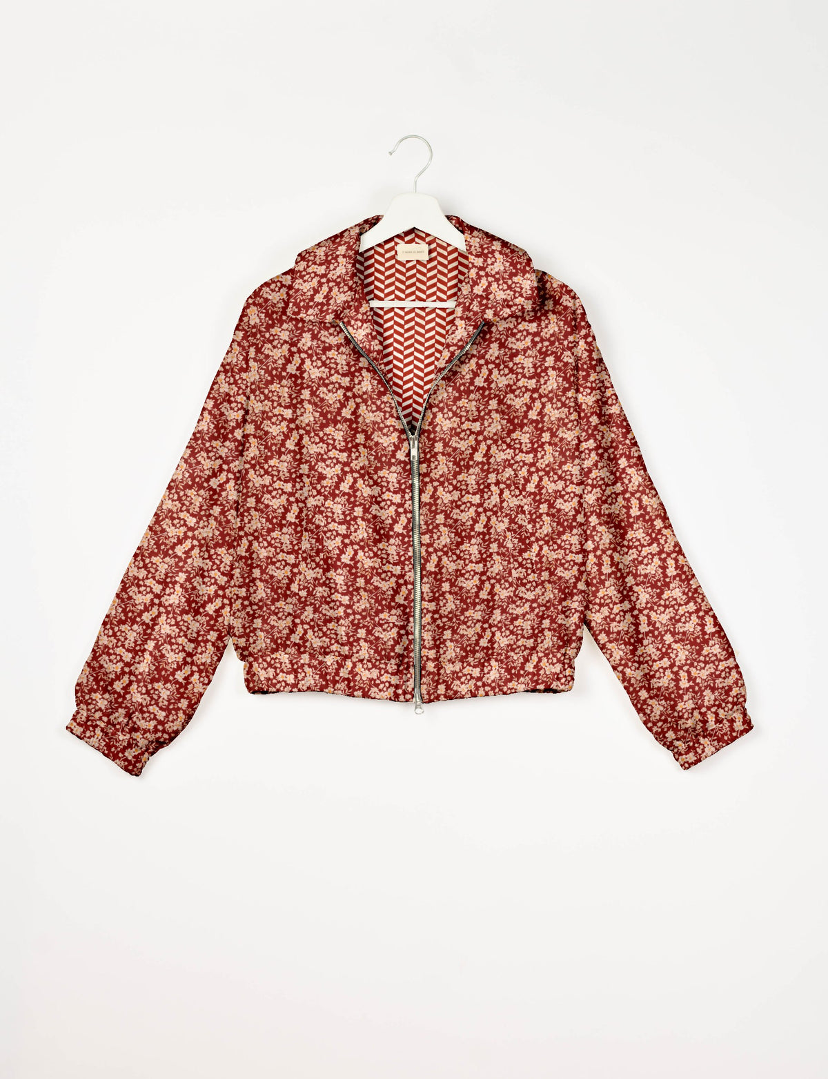 Red floral bomber jacket with front zipper, collar, elastic cuffs, and chevron patterned inner lining, hanging on white