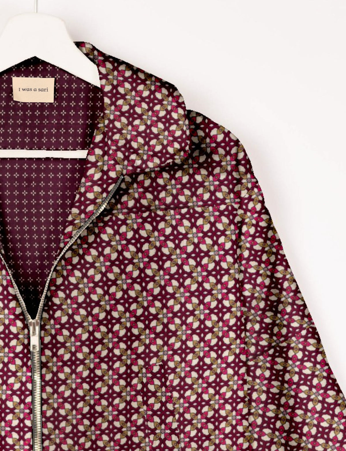 Red geometric patterned bomber jacket with silver zipper, hanging on white hanger, label reads "I was a sari" inside collar