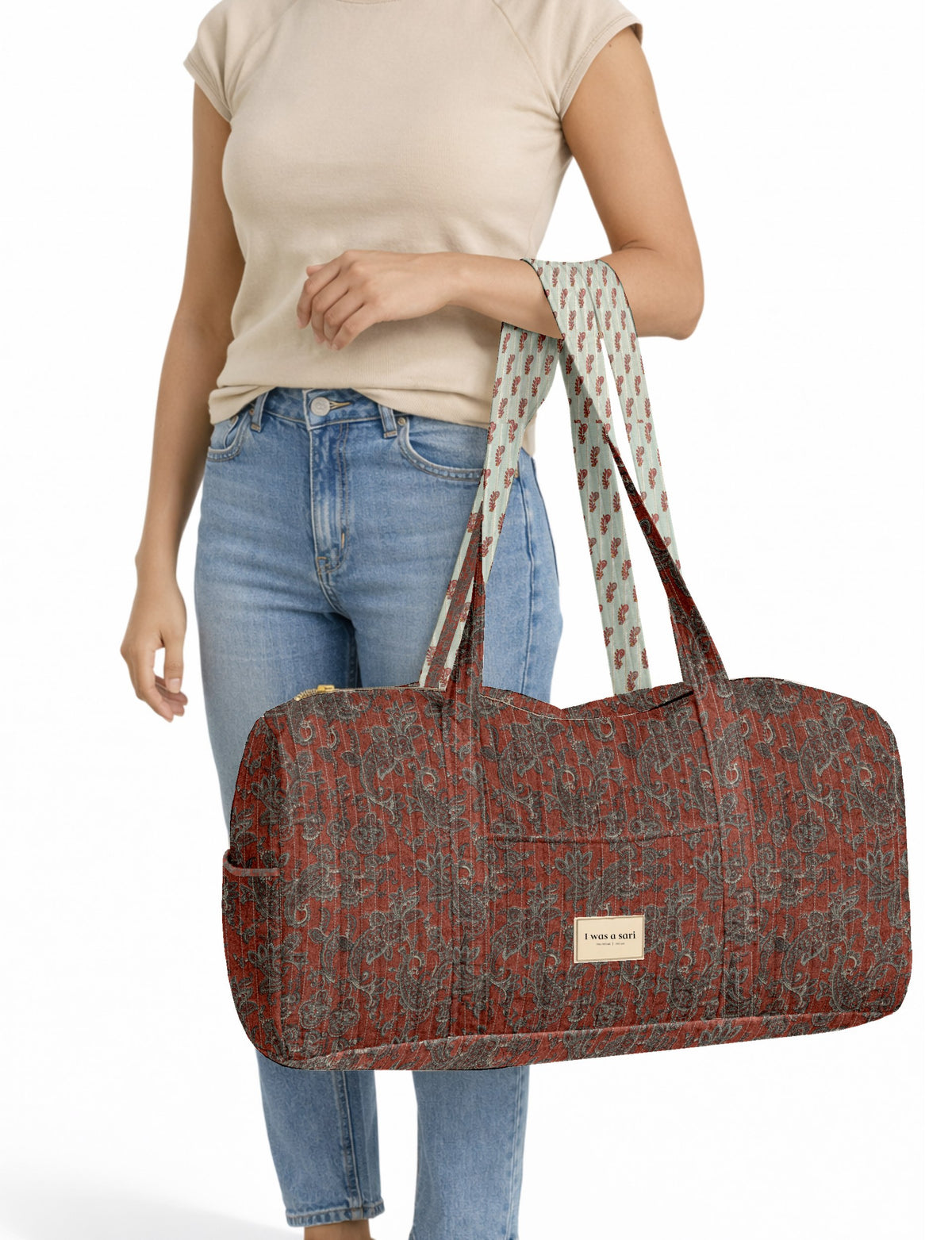 Woman wearing beige top and blue jeans holding a red paisley quilted barrel bag with patterned handles and front label