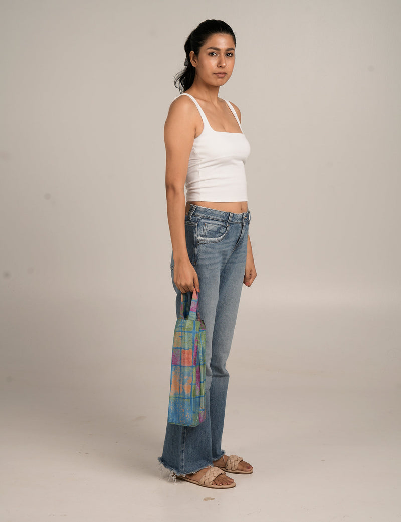 Model wearing blue paisley patterned wine bag with multicolor grid design, held by short handles, side view on plain background