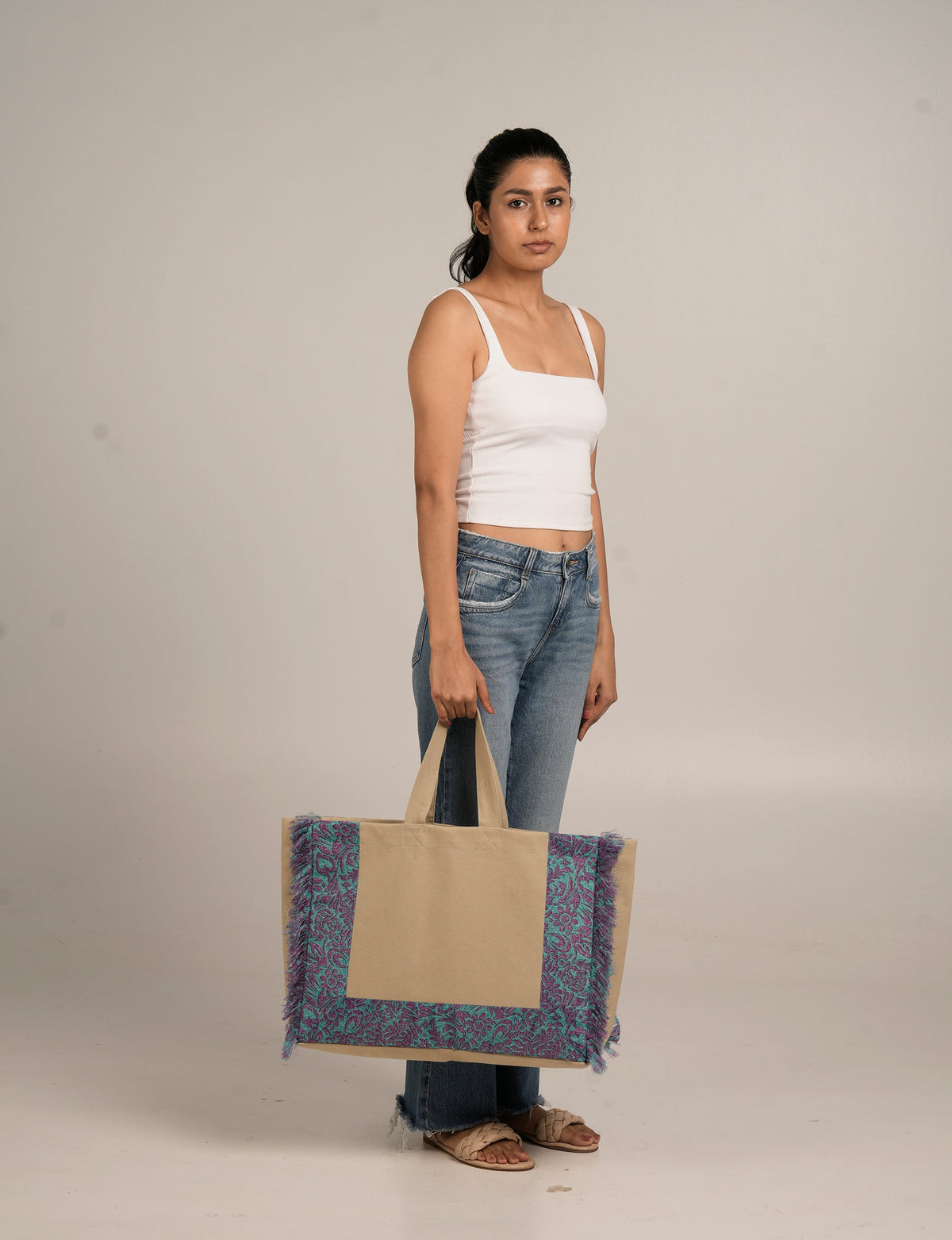 Woman wearing white tank top and blue jeans holding large beige tote bag with purple and teal floral fringe border