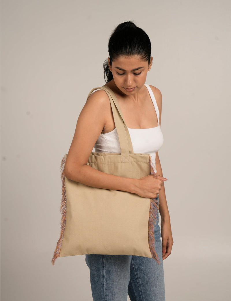 Woman wearing beige canvas tote bag with multicolor fringe on sides, shown from front and side angles, holding strap on shoulder