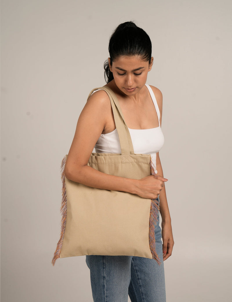 Woman wearing a beige fringe shopper bag with long handles over shoulder, paired with white tank top and blue jeans, side view