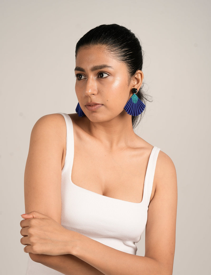 Model wearing pleated fan-shaped earrings in blue and teal with black stud, paired with white ribbed tank top, front view