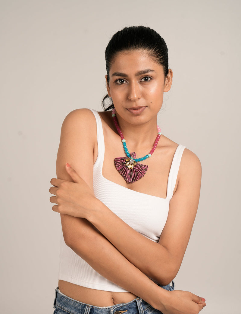 Woman wearing a pleated pink abstract necklace with blue, pink, and white beads, paired with a white tank top and denim jeans