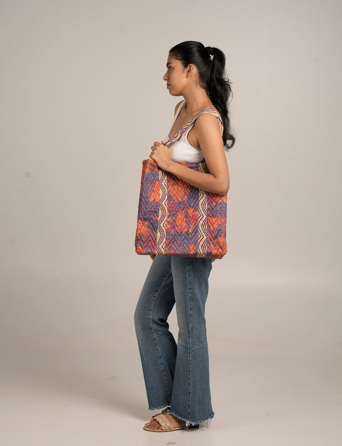 Woman wearing a sleeveless white top and blue jeans carrying a quilted tote bag with orange, purple, and beige floral patterns