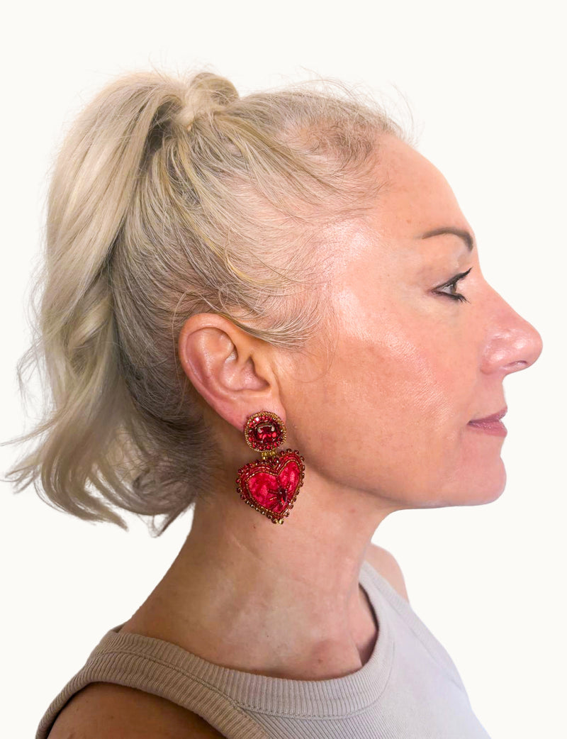 Woman wearing pink abstract heart drop earrings with red and gold bead details, side profile view on white background
