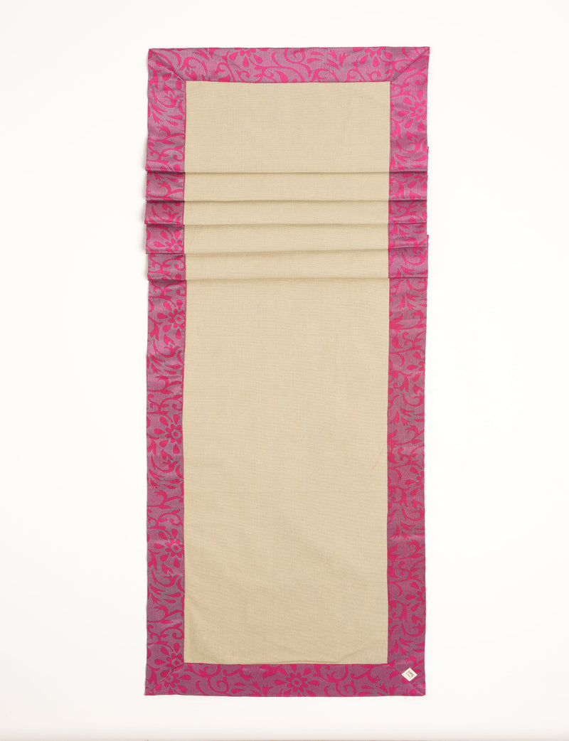 Beige table runner with pink floral patterned border, flat laid with visible fabric texture and a small corner label