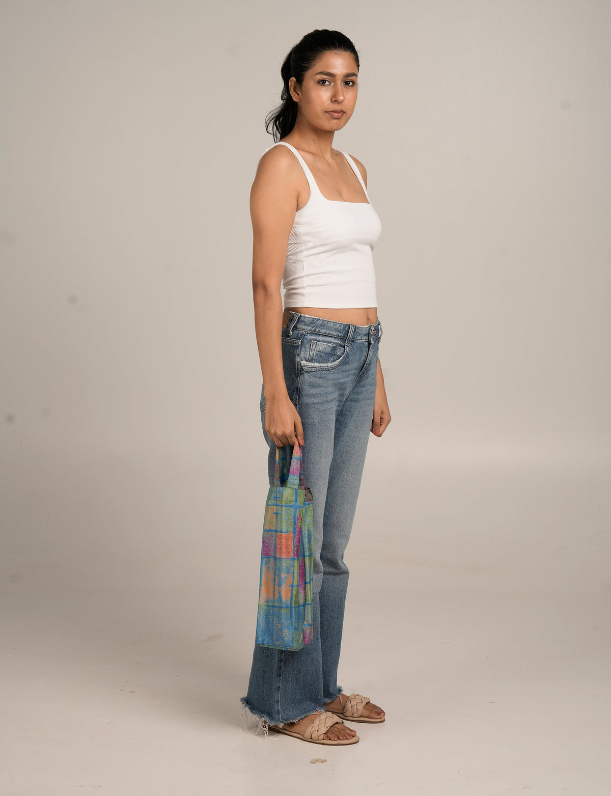 Woman wearing white tank top and blue jeans holding a multicolor patterned wine bag by its handle in side view